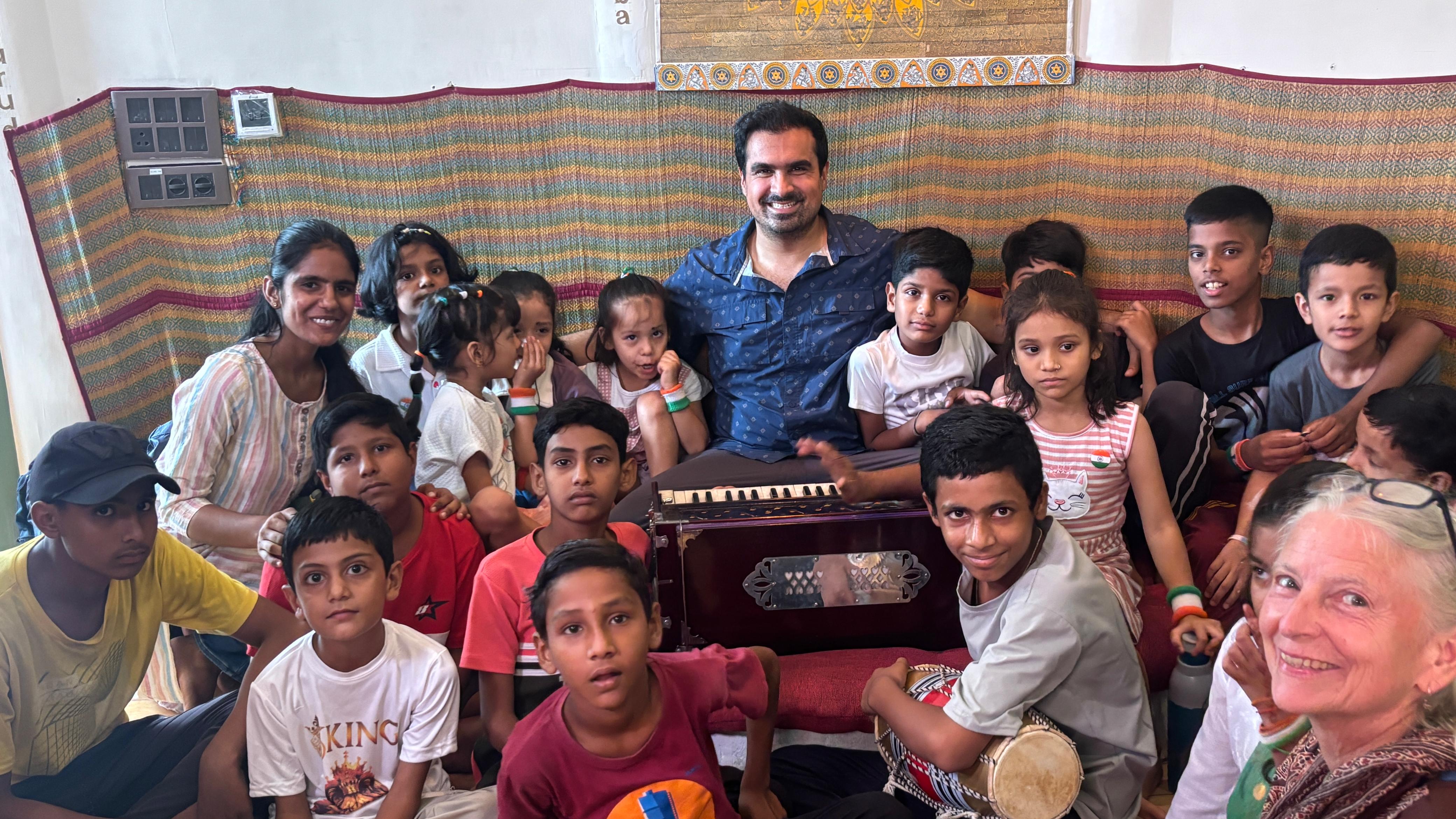 Children, teachers, and participants gathered around a harmonium after a music session